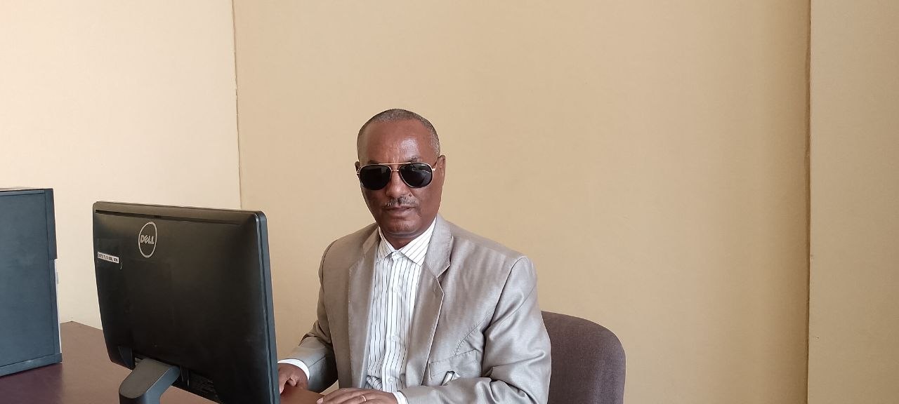 Gebrekidan Hadush, Executive Director of TDOU — a man with brown skin wearing dark sunglasses, a striped shirt, and a grey suit jacket, seated at his desk next to a computer monitor.